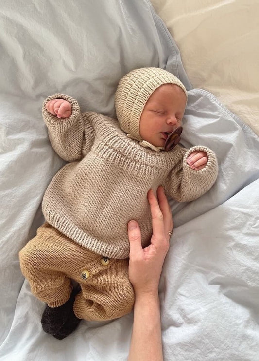 A sleeping baby lies on a soft bed in a knitted beige outfit, including a hat and pants. A hand gently rests on the baby's chest, conveying warmth and care.