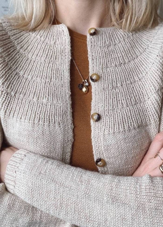 A person wearing a textured beige cardigan with large buttons over a brown shirt. They have blonde hair and their arms are crossed, conveying a cozy vibe.