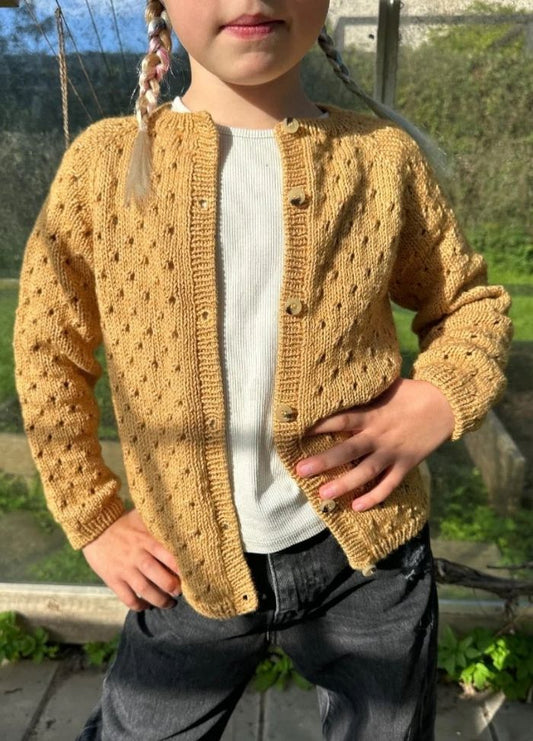 A child with braided hair wears a textured yellow cardigan over a white shirt. They stand confidently with hands on hips, sunlight casting shadows.