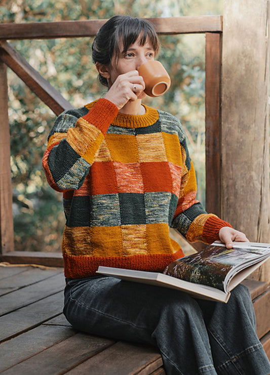 Person wearing the Berta Sweater, a colorful checkered knit in orange, yellow, green, and red, seated outdoors with a book and mug.