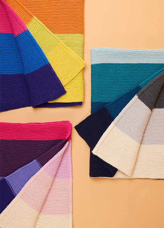Four hand-knitted Brighton Blankets arranged in a square on a light brown background – top left with rainbow stripes, top right in blue and gray tones, bottom left in pink and lavender hues, and bottom right in cream, beige, navy, and teal stripes.