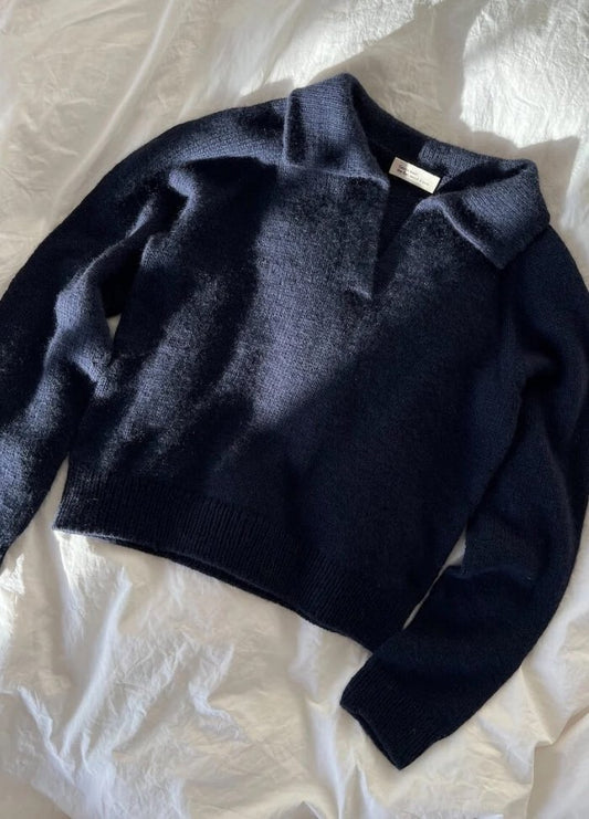 A dark navy, long-sleeve sweater with a V-neck and collar, is spread on a textured white fabric. Sunlight casts soft shadows, creating a cozy ambiance.