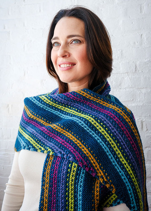 Person wearing Esme’s Sister Shawl with horizontal stripes in blue, green, yellow, pink, and purple, standing against white brick wall.