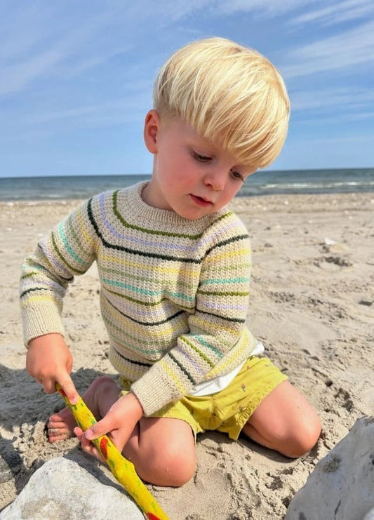Young child with blond hair plays in the sand at a beach, wearing a striped sweater and yellow shorts. The scene feels serene and focused.