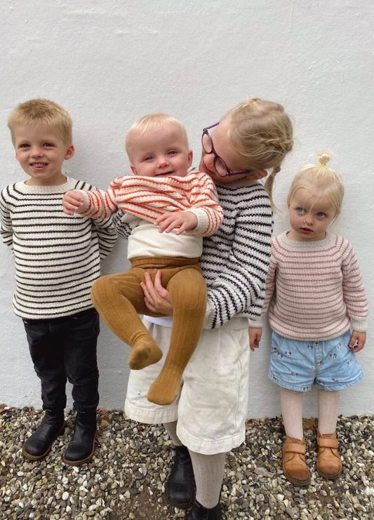 Four children stand against a white wall, all wearing striped sweaters. One older child holds a baby, and the scene conveys a warm, familial connection.