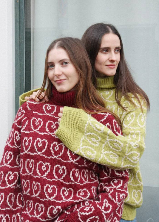 Two people stand close, smiling warmly. One wears a red sweater, and the other a green sweater, both with white heart patterns, conveying friendship.