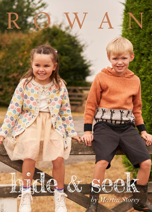 Two children, a girl and a boy, sit smiling on a wooden fence. The girl wears a patterned cardigan and tulle skirt; the boy dons an orange sweater. Lush greenery surrounds them.
