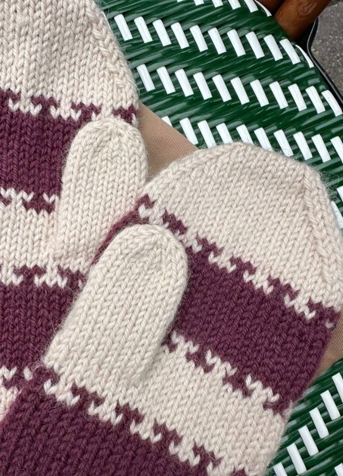 Knitted mittens with beige and maroon stripes rest on a green and white woven surface.