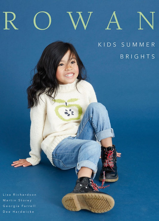 Smiling child sits against a blue background, wearing a white sweater with a green apple design, rolled-up jeans, and black boots. Text reads "Rowan Kids Summer Brights."