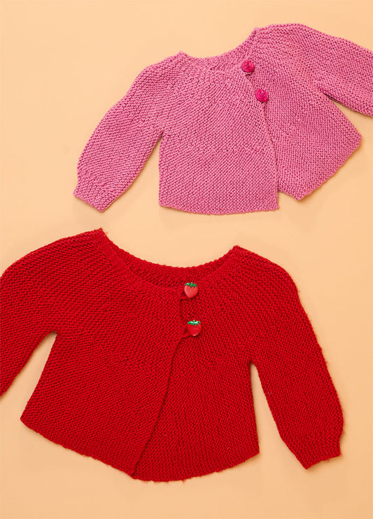 Flat lay of two hand-knitted baby cardigans on a peach background – pink cardigan with matching buttons and red cardigan featuring strawberry-shaped buttons, perfect for cozy and charming babywear.