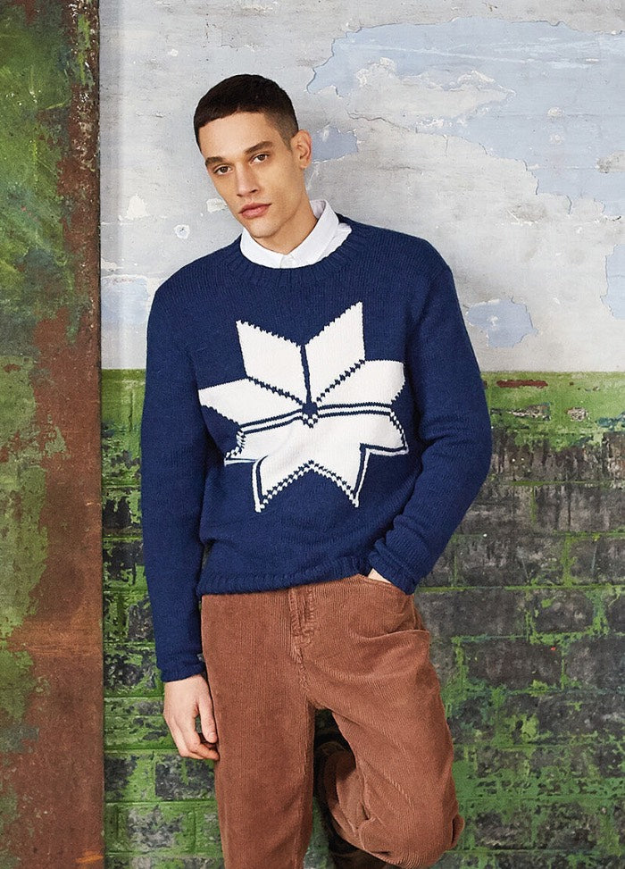 A man stands against a textured wall, wearing a blue sweater with a large white geometric star and brown corduroy pants, conveying a casual tone.