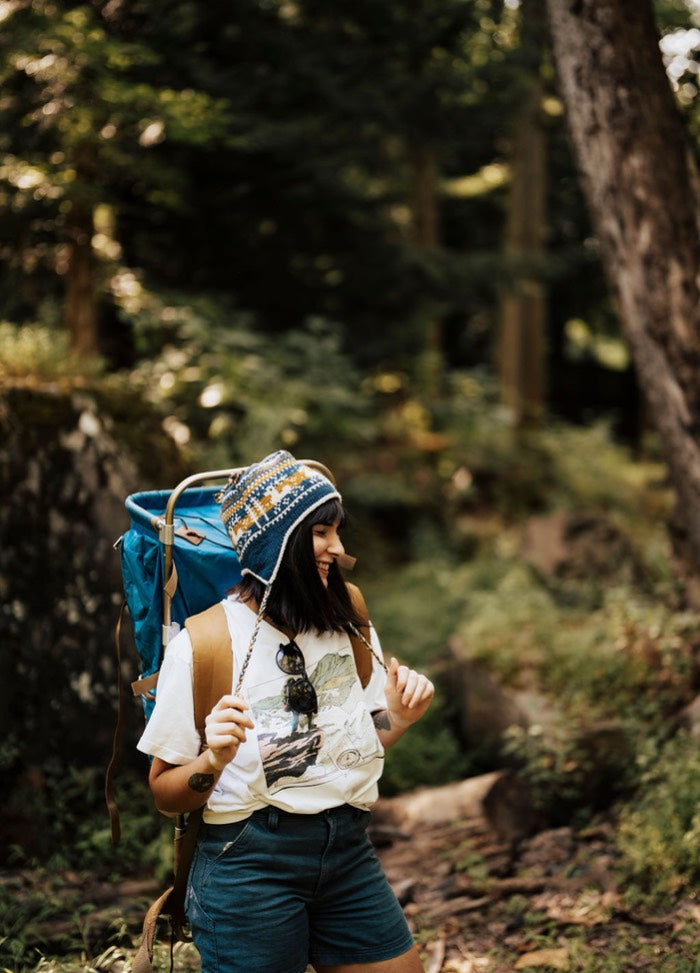 Colorful earflap hat worn with a graphic tee and blue shorts, hiking with a large blue backpack in the forest.
