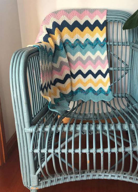 Miami Baby Blanket – cheerful knitted zigzag pattern in pink, yellow, white, teal, and navy blue, draped over a light blue wicker chair for a playful and cozy nursery accent.