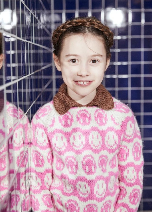 Young girl with braided hair smiles warmly, wearing a pink sweater with smiley faces and brown collar. Reflected in blue tiled wall, evoking cheerful vibe.