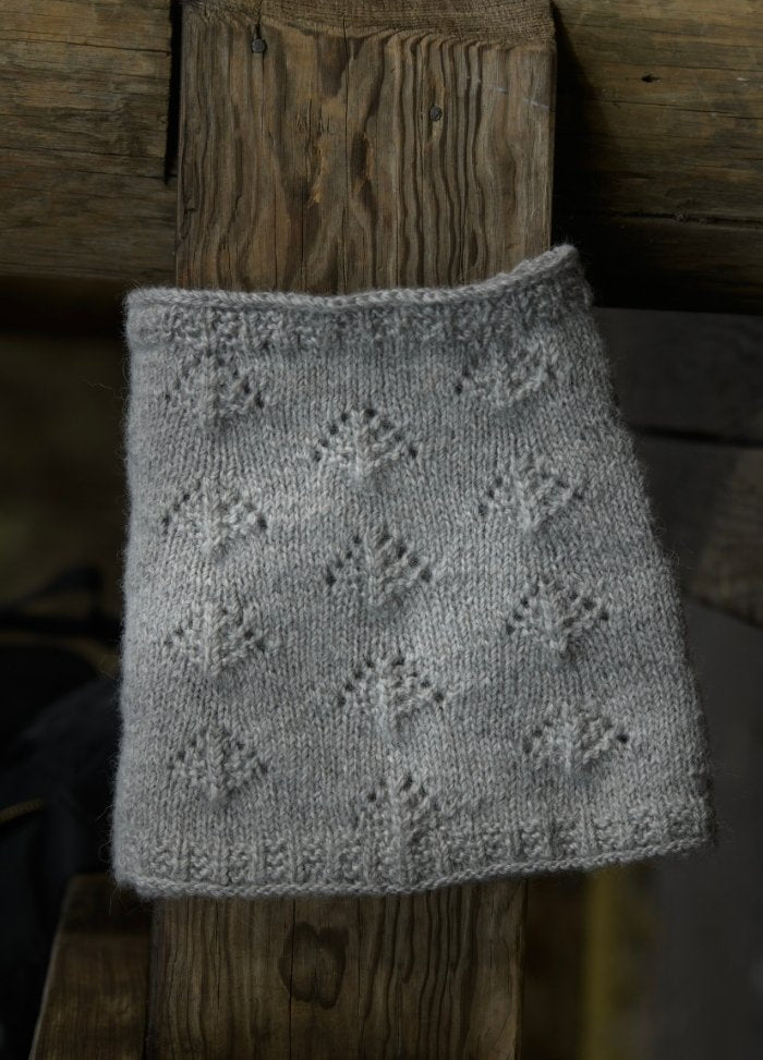 A gray knitted fabric draped over a rustic wooden surface, featuring intricate diamond and leaf patterns. The setting conveys a cozy, handcrafted feel.