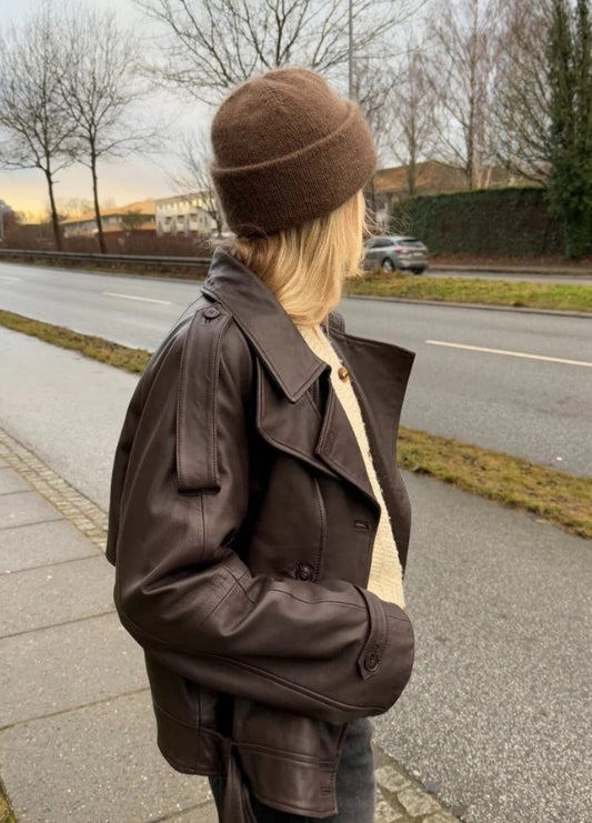 A person stands on a sidewalk facing away, wearing a brown beanie and dark leather jacket. It's a cloudy day with bare trees lining the road.