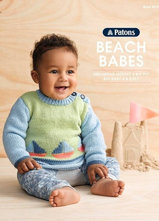A cheerful baby wearing a pastel green and blue knitted sweater with boat designs sits on a light wooden floor, next to a sandcastle. The text reads “Patons Beach Babes.”