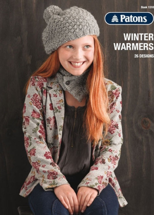 A smiling girl with long red hair wears a knitted gray hat and scarf, floral jacket, and dark blouse. The text reads: "Patons Winter Warmers, 26 Designs." The tone is cozy and cheerful.