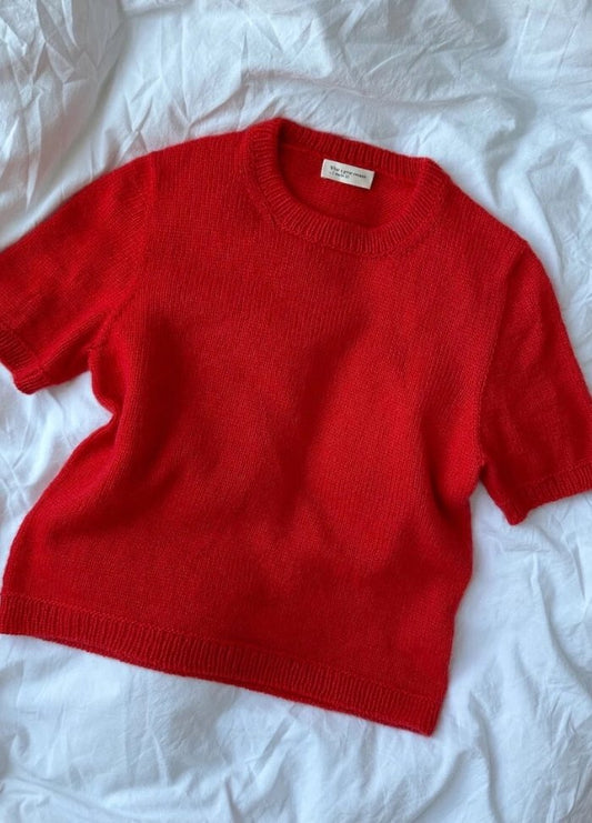 A vibrant red knitted sweater with short sleeves is laid flat on a soft, crumpled white fabric background, contrasting warmly with its surroundings.