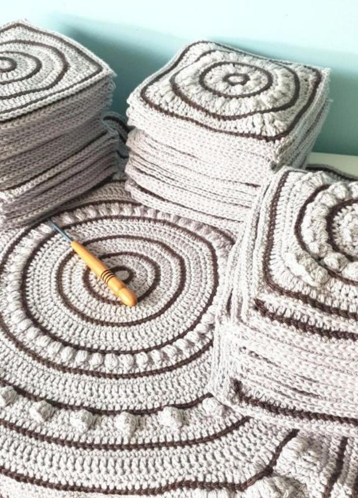 Stacks of crocheted beige and brown squares with spiral patterns, neatly arranged. A wooden crochet hook rests on one piece, suggesting craft and creativity.
