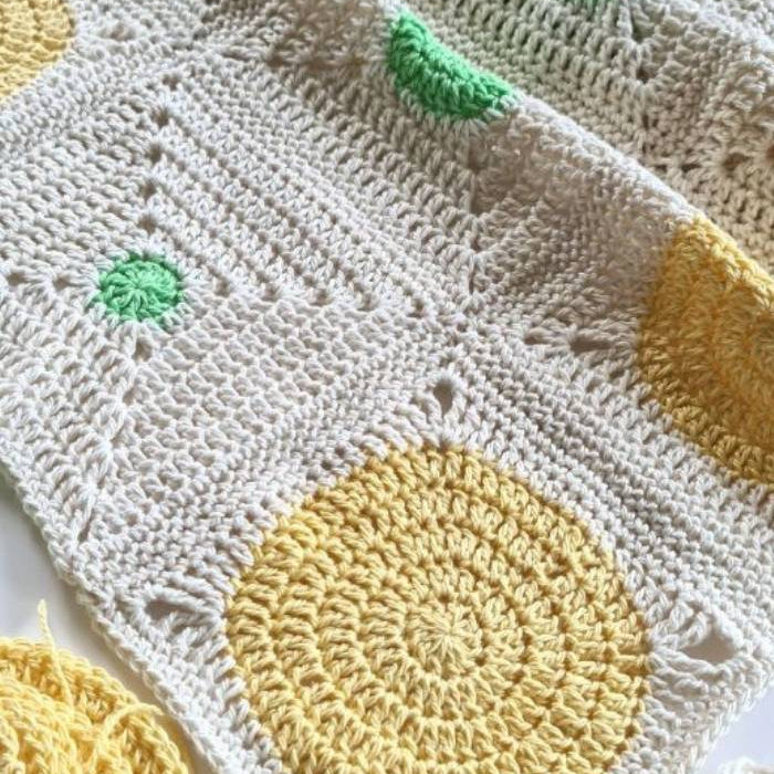 Crocheted blanket with circular patterns in yellow and green on a cream background. Yarn balls are visible, suggesting a warm, creative atmosphere.