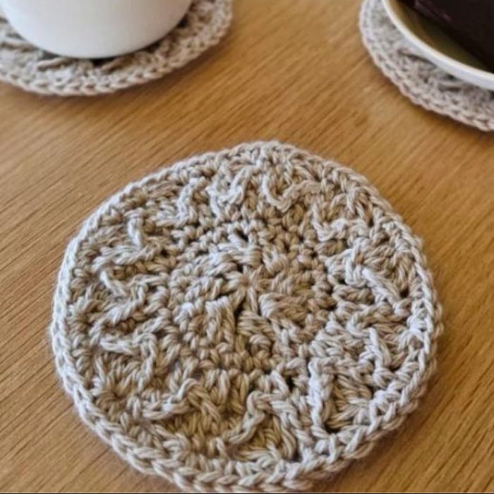 A white mug of tea and plate with chocolate sit on a wooden table, each on a beige crocheted coaster. Another coaster is in the foreground. Cozy and inviting.