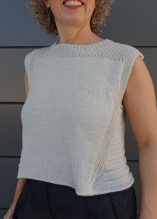 Woman wearing a sleeveless, light-colored knitted Summer Esio Top with ribbed neckline and armholes, styled with dark pants. Lightweight yarn and minimalist design ideal for warm weather fashion.