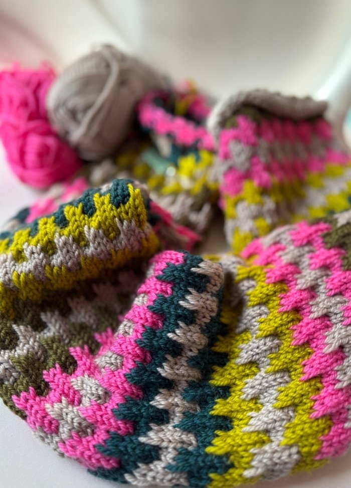 Close-up of colorful crochet work with bright pink, yellow, gray, and green stitches. Yarn balls in the background, creating a cozy, vibrant feel.