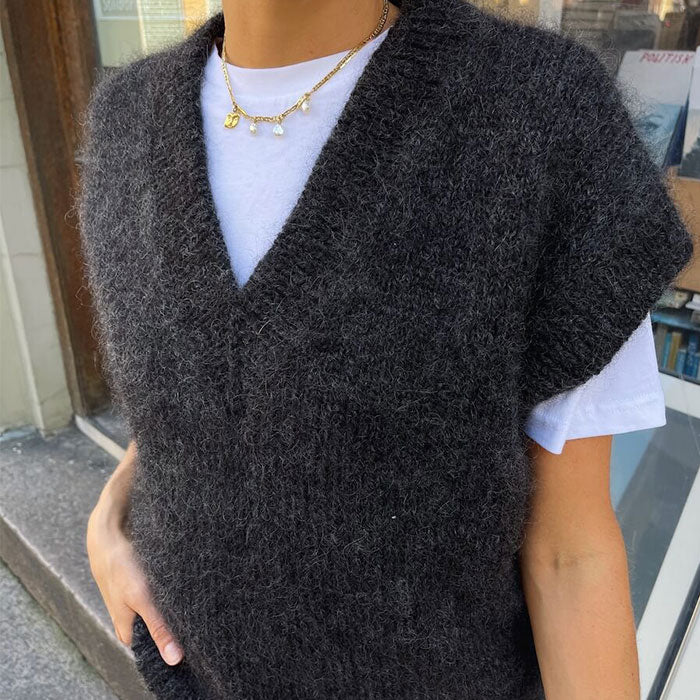 A person wearing a dark grey, fuzzy sleeveless sweater over a white T-shirt stands outdoors. They hold a beige textured bag, exuding a cozy, casual vibe.