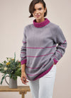 Smiling person in a purple sweater with pink stripes and collar, hands in white pants pockets. A bouquet in a jug sits on a wooden table nearby.