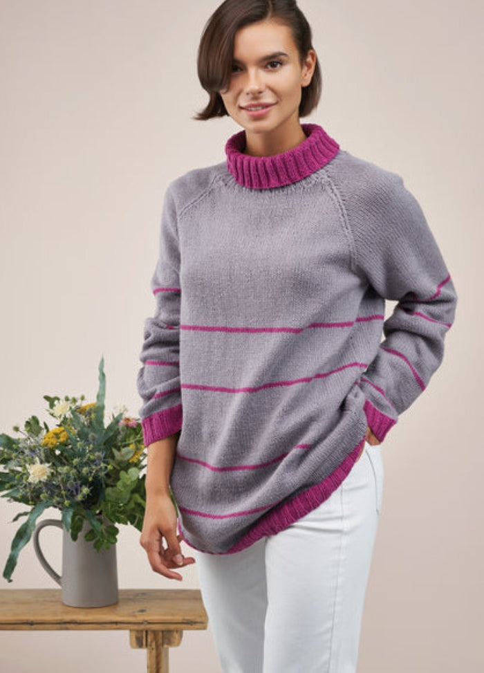 Smiling person in a purple sweater with pink stripes and collar, hands in white pants pockets. A bouquet in a jug sits on a wooden table nearby.