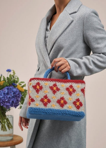 A woman holds a colorful knitted bag adorned with flowers, showcasing her unique style and love for handmade accessories.
