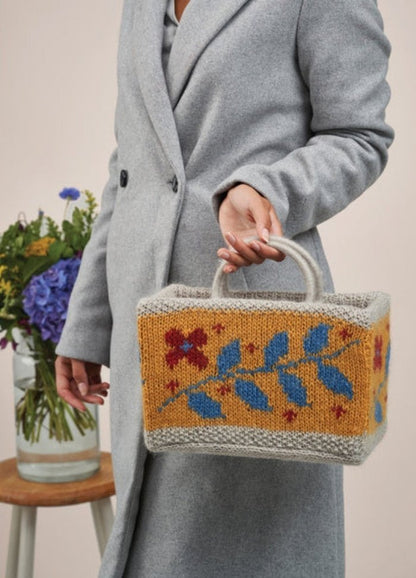 A woman displays a vibrant knitted bag featuring floral designs, highlighting her fashion sense and appreciation for crafts.