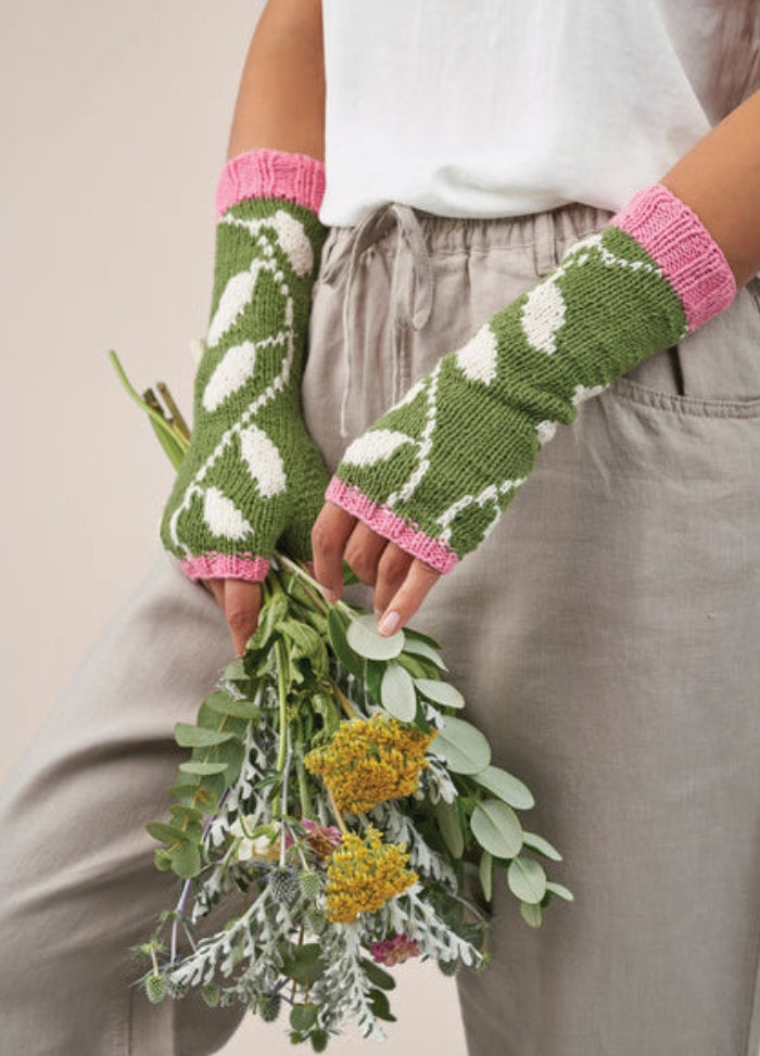 A person wearing green knit fingerless gloves with white leaf patterns and pink cuffs holds a colorful bouquet of flowers. They are dressed casually.