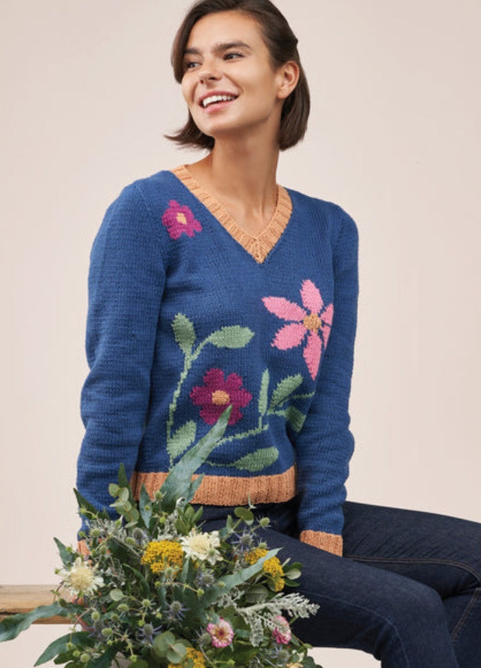 Smiling woman sitting, wearing a blue sweater with bright floral patterns and jeans. She holds a bouquet of wildflowers, exuding a cheerful and cozy vibe.
