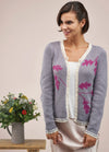 Smiling woman wearing a lavender cardigan with pink leaf patterns and cream trim, standing next to a table with a jug of colorful flowers. Serene atmosphere.