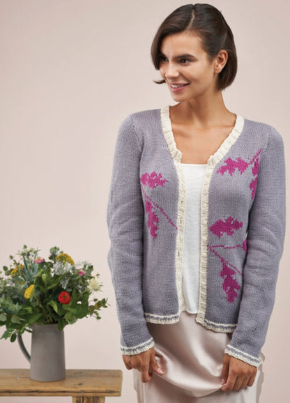 Smiling woman wearing a lavender cardigan with pink leaf patterns and cream trim, standing next to a table with a jug of colorful flowers. Serene atmosphere.