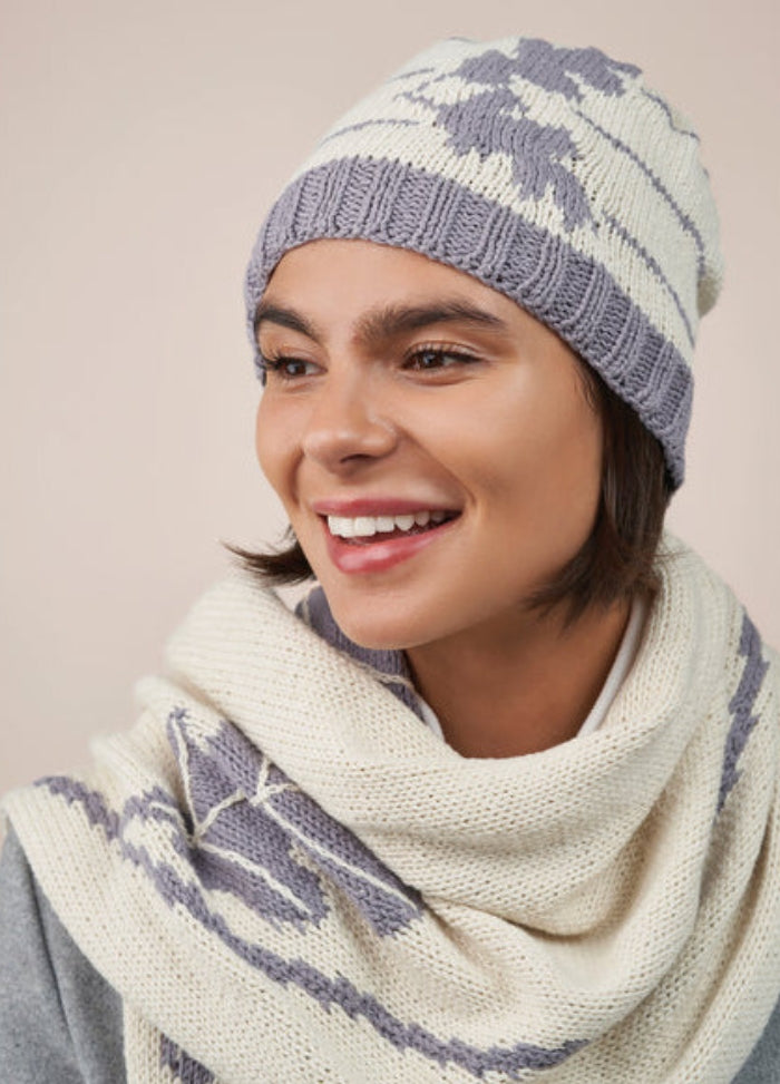 Smiling person wearing a knitted hat and scarf in a light lavender and cream pattern. The background is a soft beige, giving a cozy vibe.