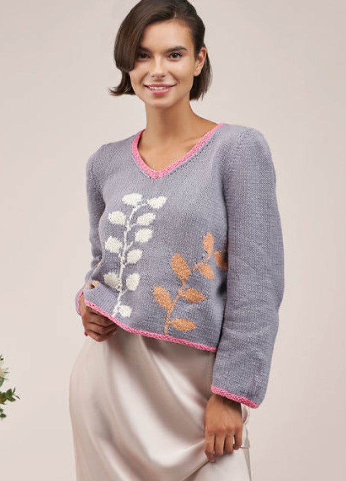 Smiling woman wearing a gray sweater with pink trim and leaf patterns in white and brown. The setting is bright, creating a cheerful tone.