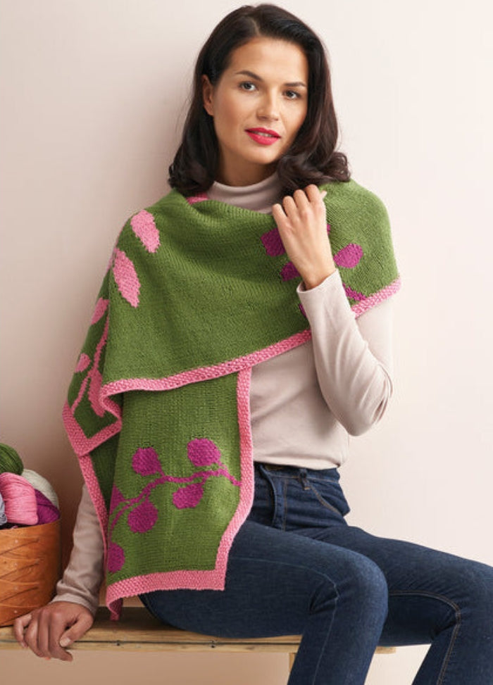 A woman sits on a bench wearing a green knit shawl with pink leaf patterns and edges. Her expression is calm, set against a neutral background.