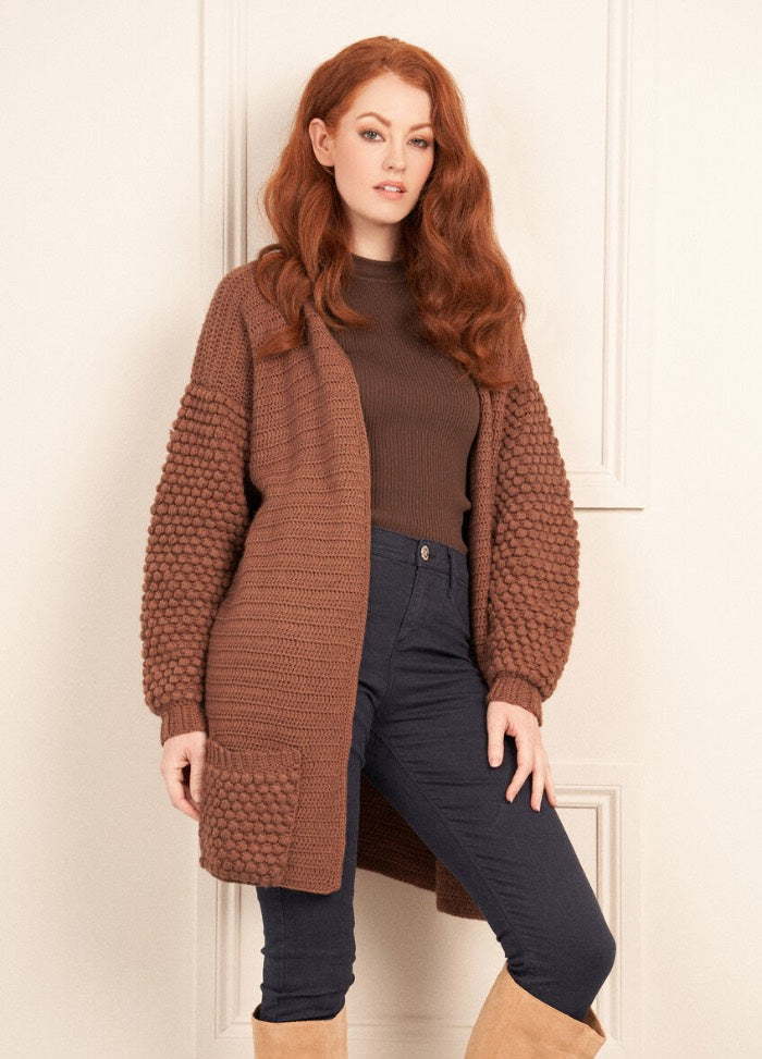 A woman with long red hair wears a brown knitted cardigan over a brown top and dark pants, standing indoors against a light-colored wall.