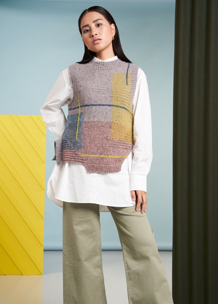A woman stands confidently, wearing an oversized white shirt and a textured, multicolored knitted vest. The background features blue and yellow panels.