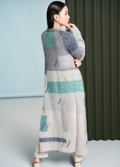 A woman in a long, patterned wool cardigan with pastel shades stands in profile, exuding elegance. The background features textured green and blue panels.
