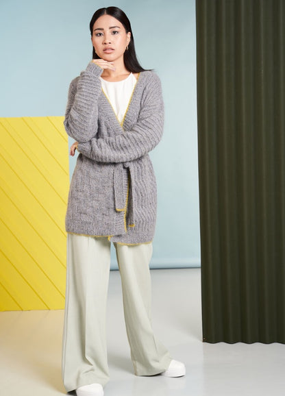 A woman stands confidently against a colorful backdrop, wearing a cozy gray cardigan and light pants. She exudes a calm, thoughtful demeanor.