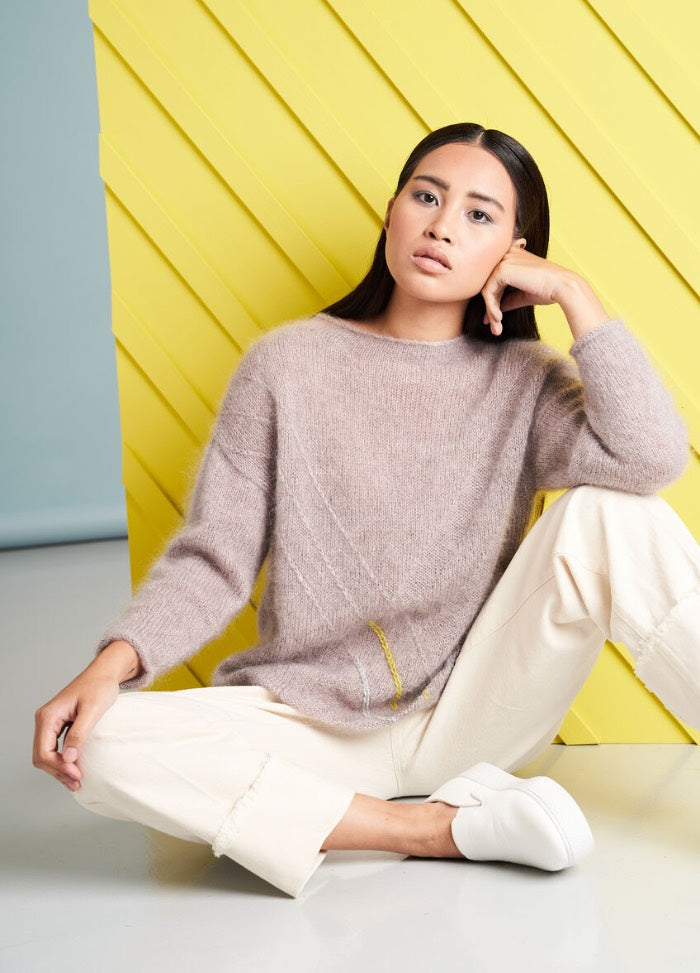 A woman with a contemplative expression sits in front of a yellow diagonal panel. She wears a cozy beige sweater, cream pants, and white sneakers.