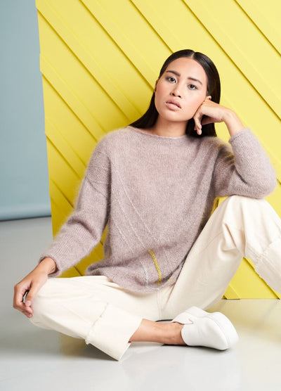 A woman with a contemplative expression sits in front of a yellow diagonal panel. She wears a cozy beige sweater, cream pants, and white sneakers.