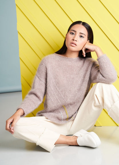 A woman with a contemplative expression sits in front of a yellow diagonal panel. She wears a cozy beige sweater, cream pants, and white sneakers.