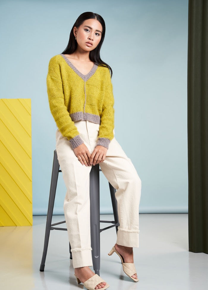 Woman sits on a stool against a blue background, wearing a yellow sweater, white pants, and beige heels. The scene appears calm and stylish.