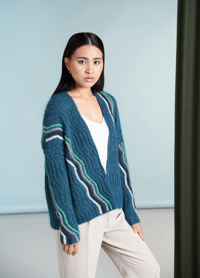 A person wearing a cozy, beige sweater with a blue and green geometric pattern and green neckline, stands against a textured green background, exuding a calm, stylish vibe.