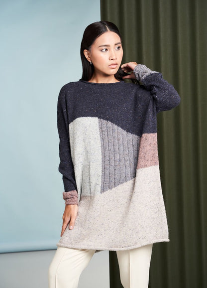 Woman in a color-block knit sweater featuring navy, cream, and pink tones poses against a blue and green backdrop, looking thoughtfully aside.